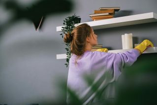 Woman in purple sweater dusts a bookshelf with candles and books.